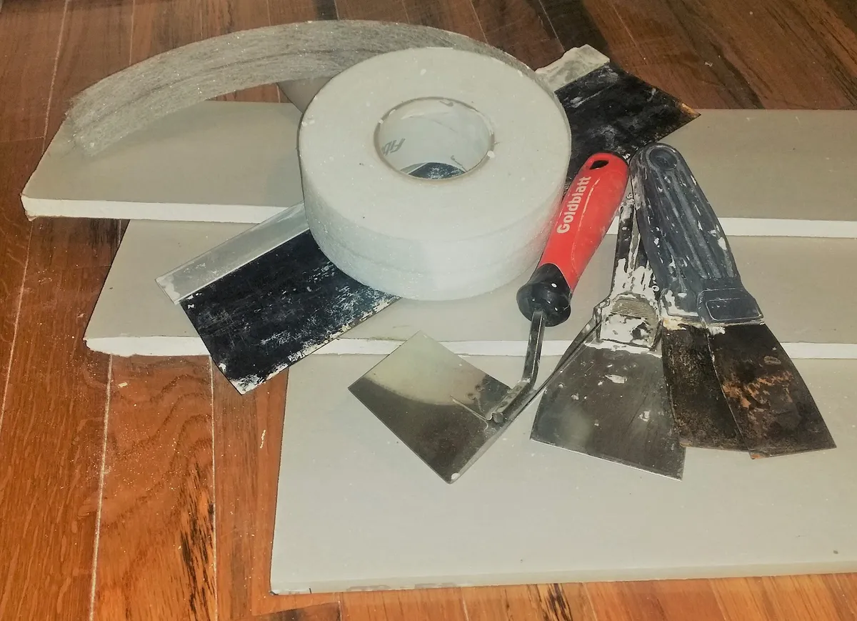 Drywall offcuts, a roll of joint tape and taping knives on a wood floor